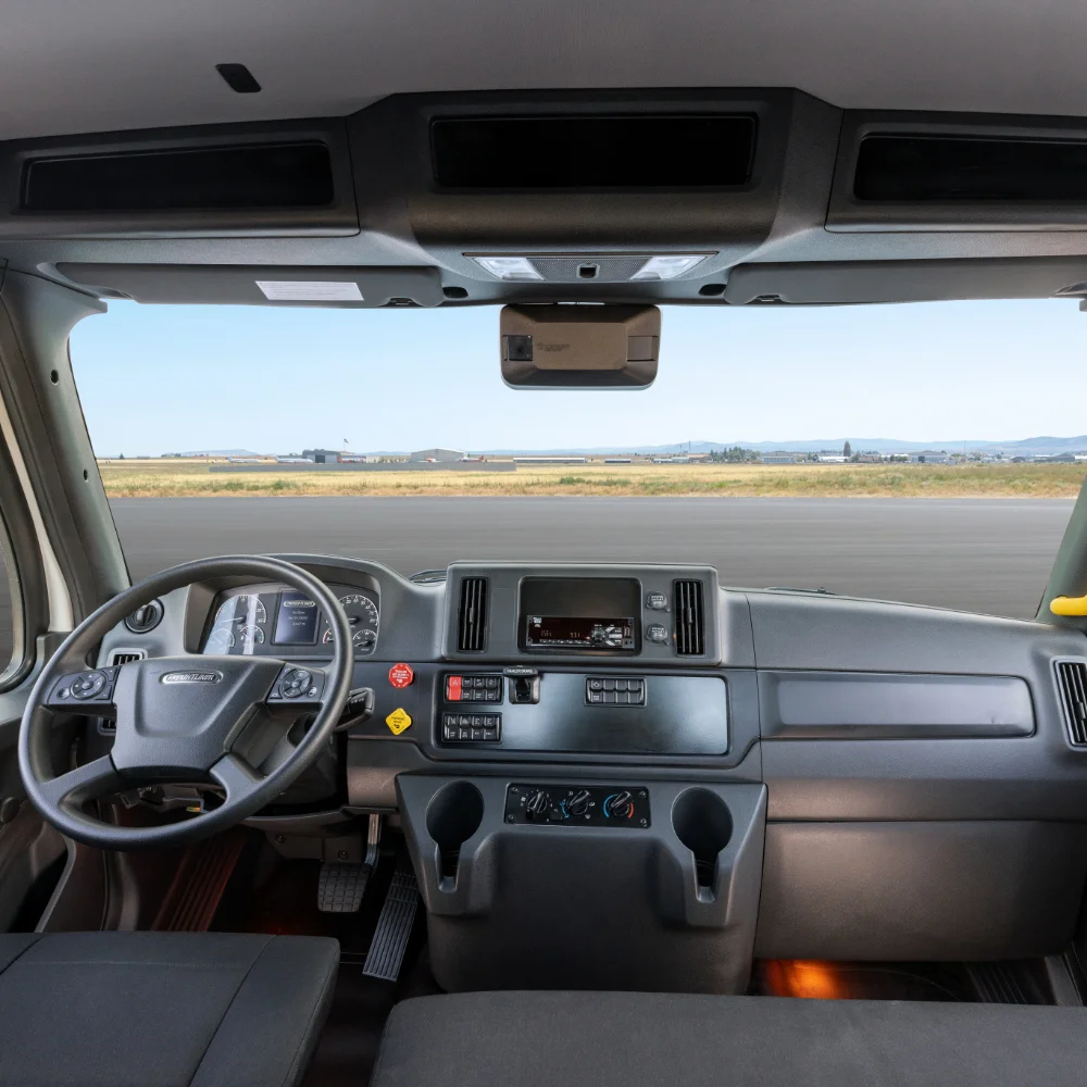 Interior view of Freightliner 114SD Plus cab highlighting the dashboard, steering wheel, and overhead storage with scenic runway view in the background.